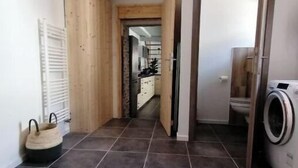 Käytävän lattia on laatoitettu, siellä on puinen vaatekaappi, pyykinpesutila, jossa on pesukone, ja taustalla näkyy keittiö.