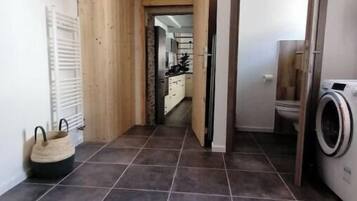 Un couloir avec un sol carrelé, une armoire en bois, un coin buanderie avec un lave-linge, et une cuisine visible au fond.