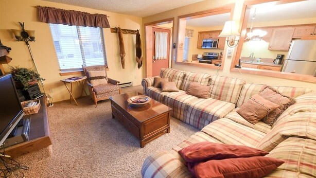 A cozy living room with a plaid sofa, a wooden coffee table, and a television. There is a chair, a lamp, and a window with curtains. A kitchen area is visible through an open door. - Trapper Condo Unit 3 (West Yellowstone)