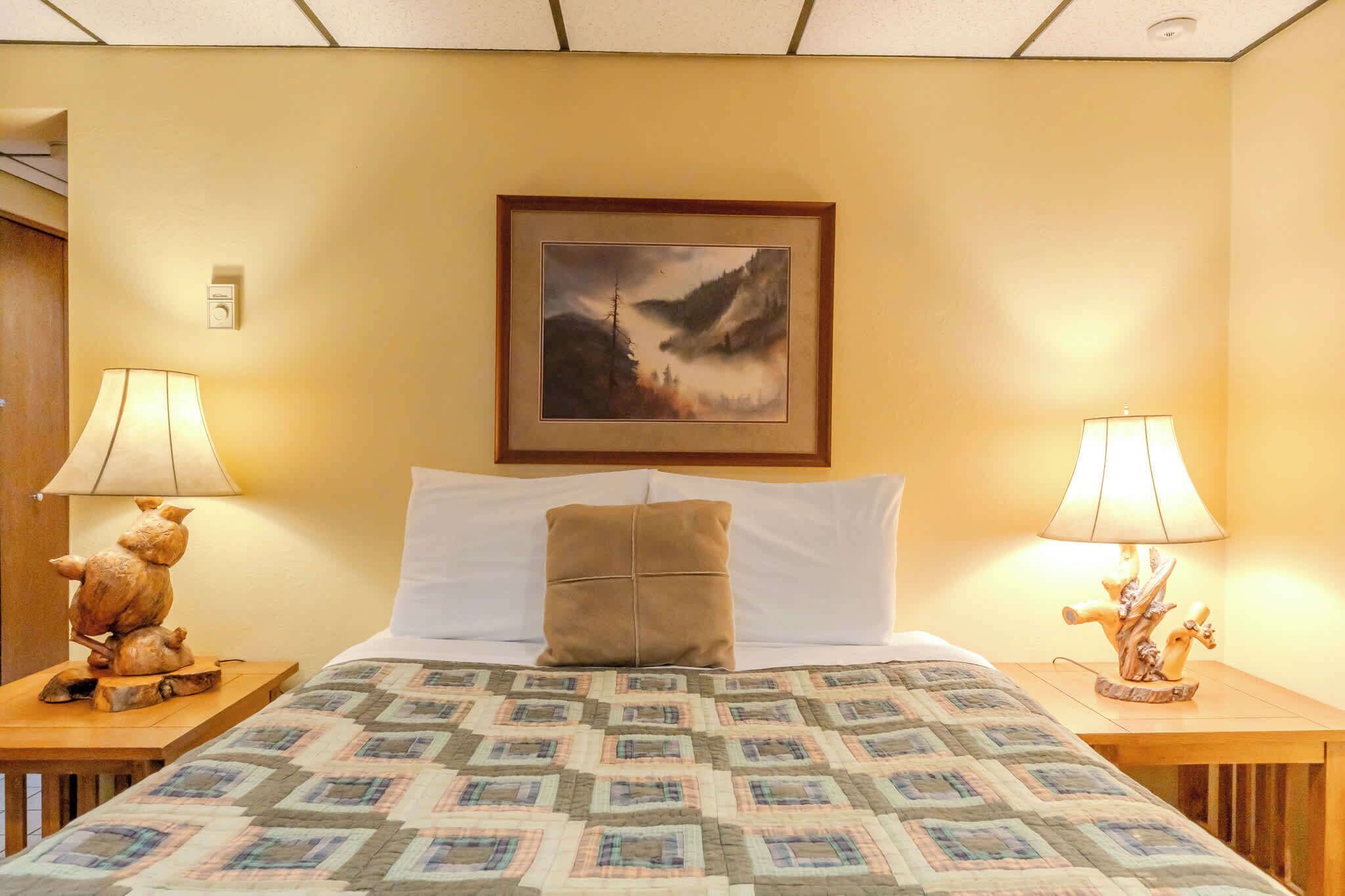 Ein Hotelzimmer mit einem Bett, zwei Nachttischen, einer Lampe, einem Gemälde und einer Holzskulptur.