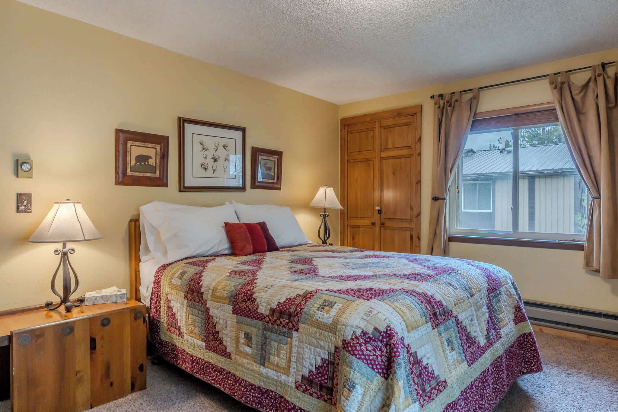 A bedroom with a bed, two bedside lamps, a wooden nightstand, a quilt, framed pictures on the wall, and a window with curtains.