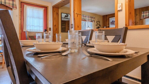 Une table à manger dressée avec des bols, des verres et des couverts, entourée de chaises en bois et agrémentée d’un rideau à carreaux.