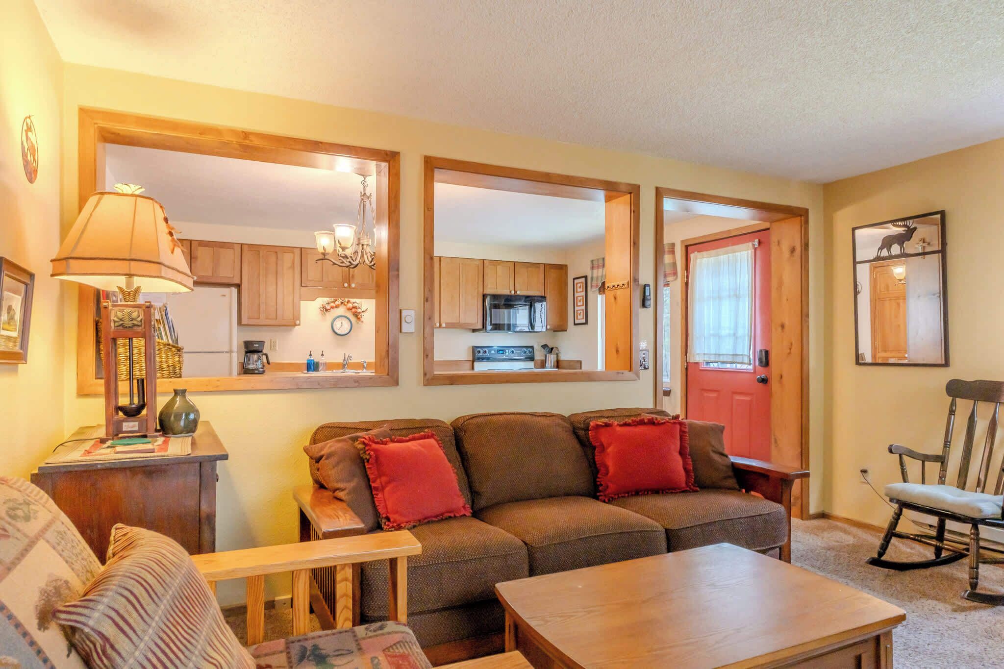 A cozy living room with a sofa, a coffee table, and a rocking chair. A kitchen is visible through an open door.