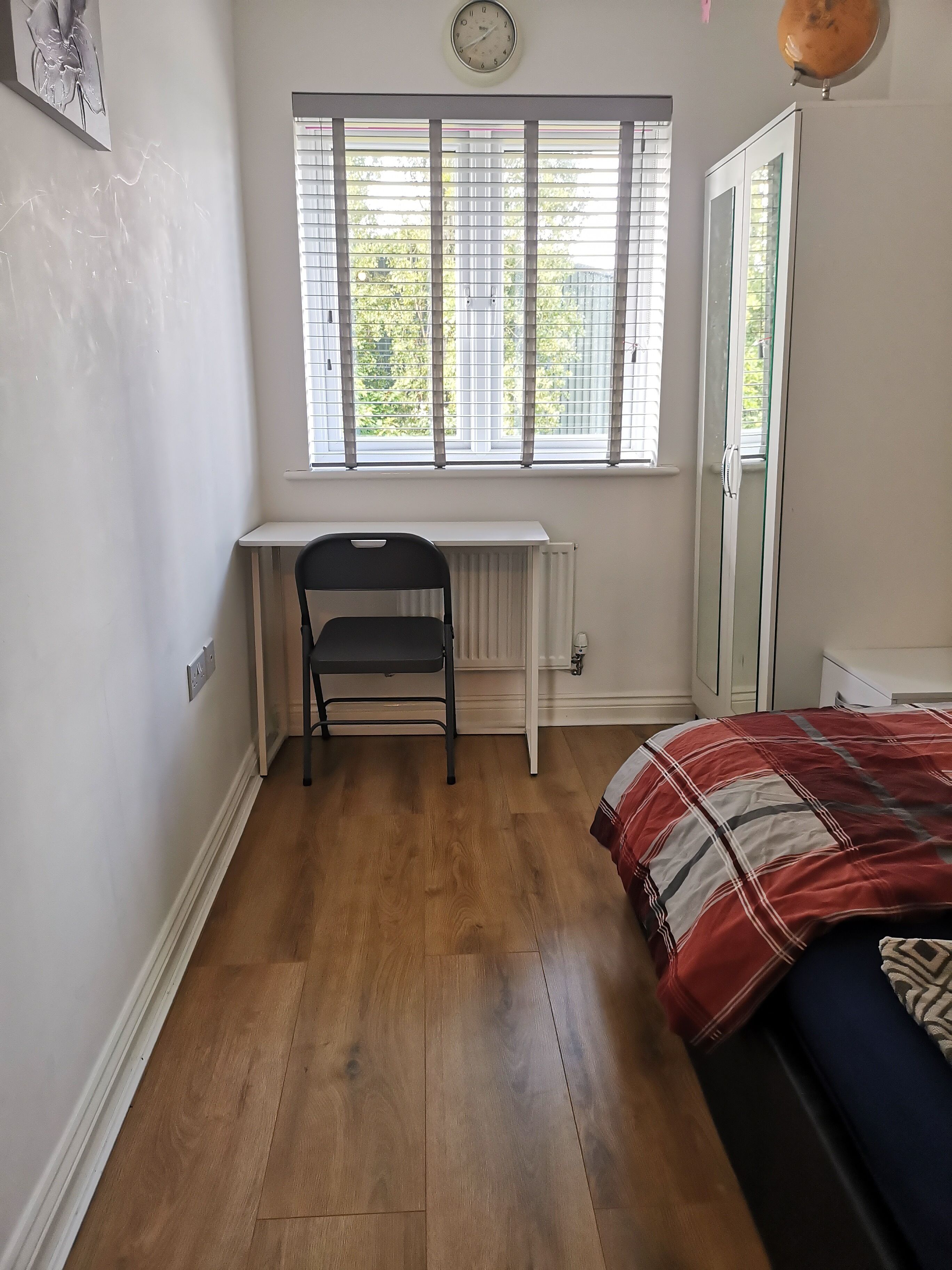 A bedroom with a wooden floor, a desk with a chair, a bed with a red and white patterned blanket, and a window with blinds.