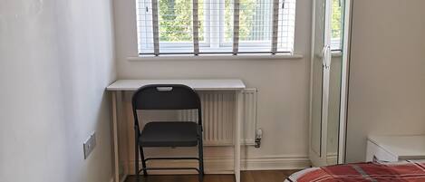 A bedroom with a wooden floor, a desk with a chair, a bed with a red and white patterned blanket, and a window with blinds.