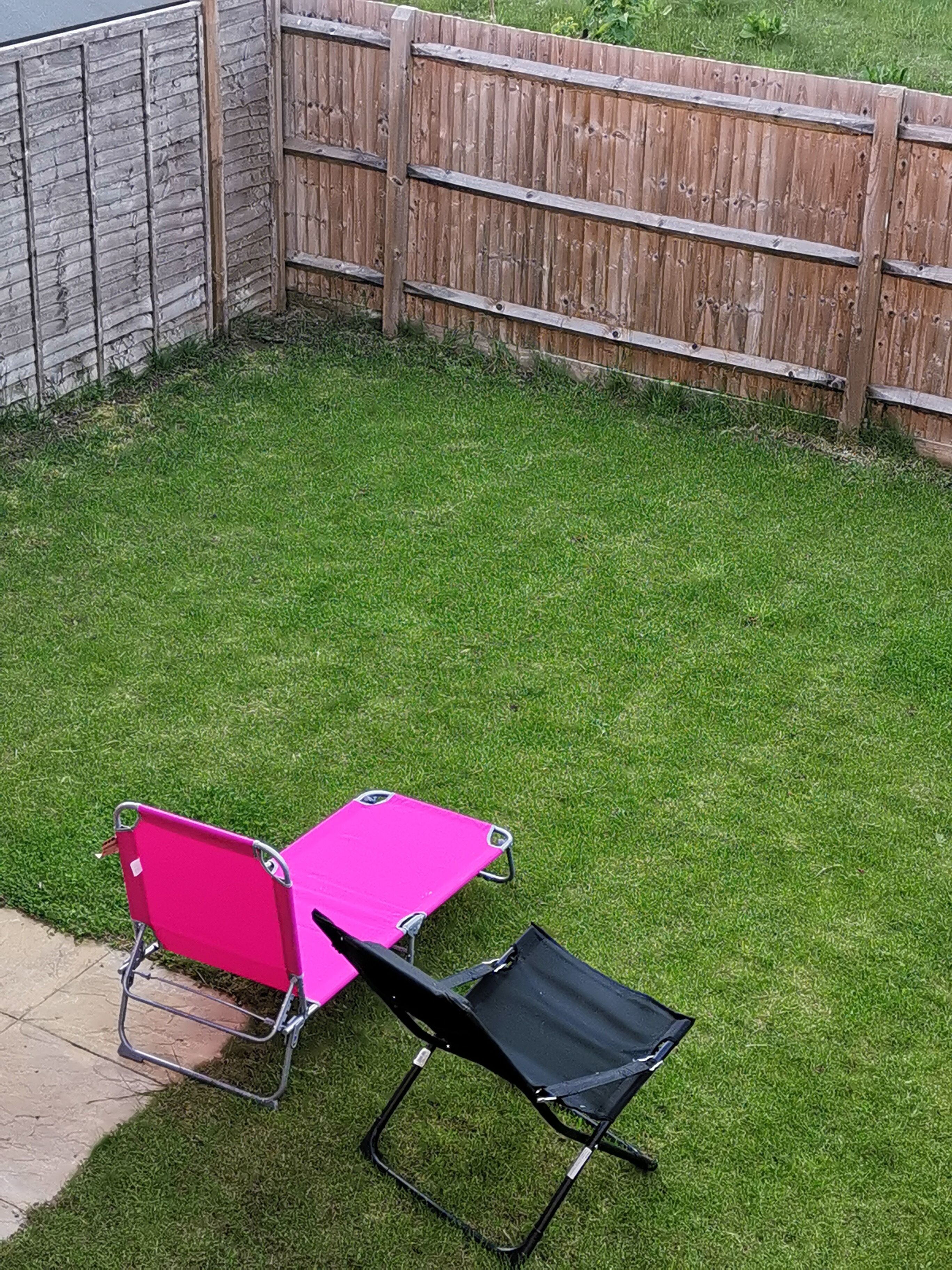 A backyard with a wooden fence, a grassy lawn, and two chairs, one pink and one black.