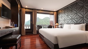 A hotel room with a large bed, a desk with a flat-screen TV, a bench, and a view of a mountainous landscape. - Aqua Of The Seas Cruise Ha Long (Tuan Chau)
