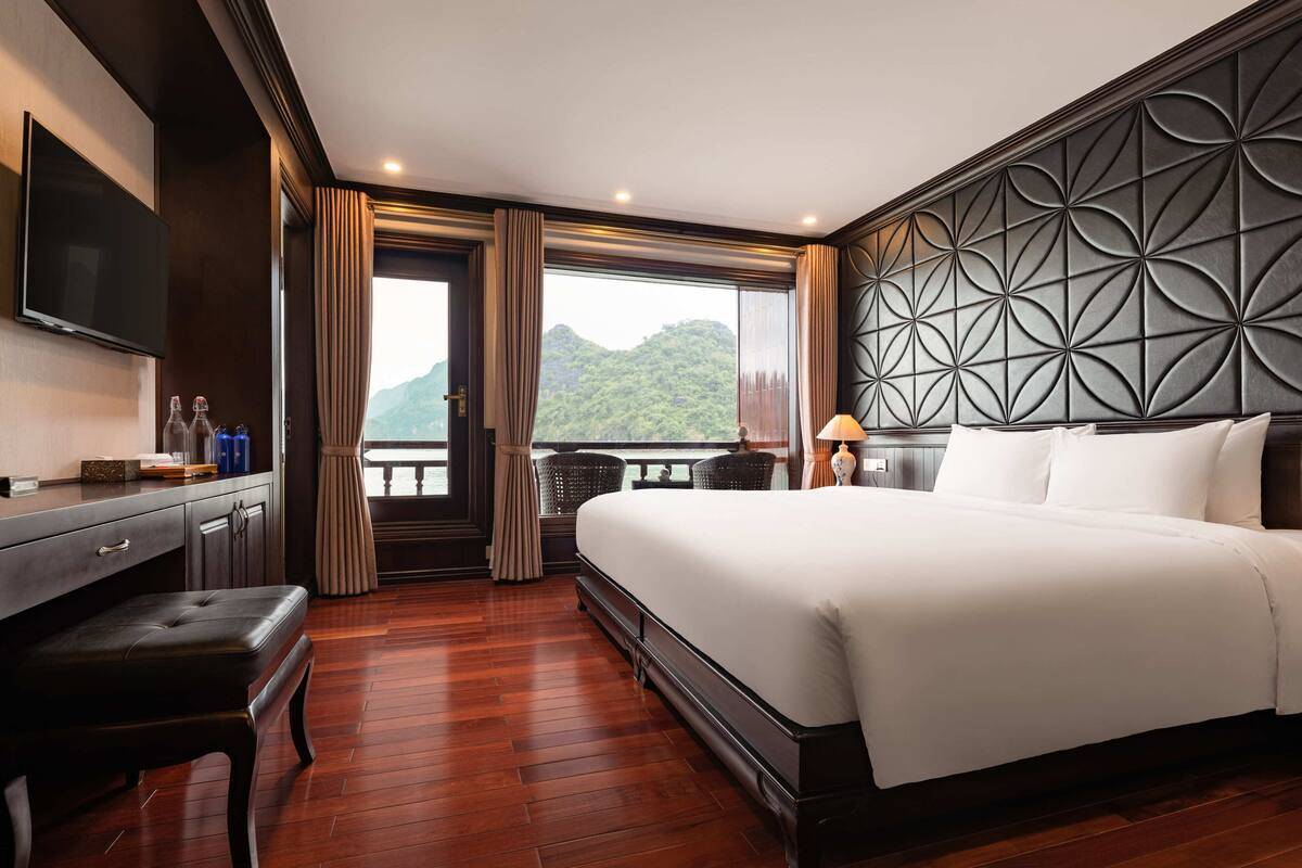 A hotel room with a large bed, a desk with a flat-screen TV, a bench, and a view of a mountainous landscape.