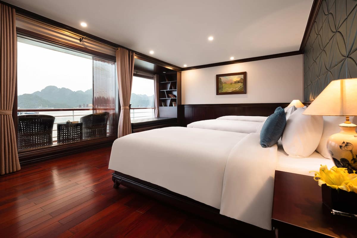 A hotel room with a large bed, a nightstand with a lamp, a bookshelf, and a view of the sea.