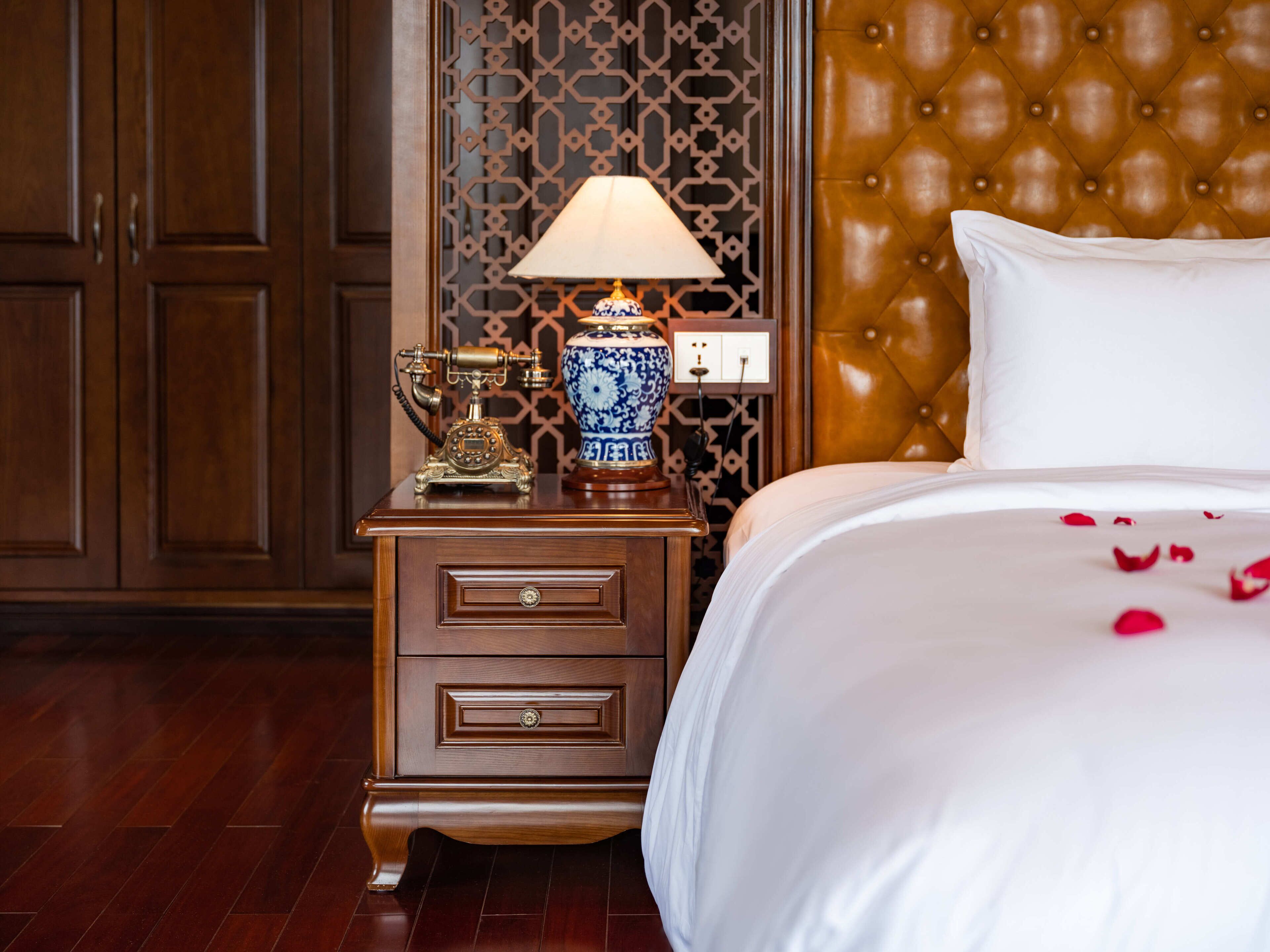 Habitación de hotel con mesita de noche de madera, teléfono antiguo, jarra azul y blanca, cama con ropa de cama blanca y cabecera capitonada.