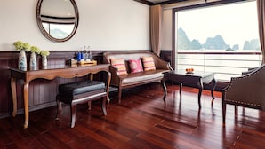 A room with a wooden bench, a sofa, a table, and a mirror. There is a window with a view of mountains and water. - Aqua Of The Seas Cruise Ha Long (Tuan Chau)