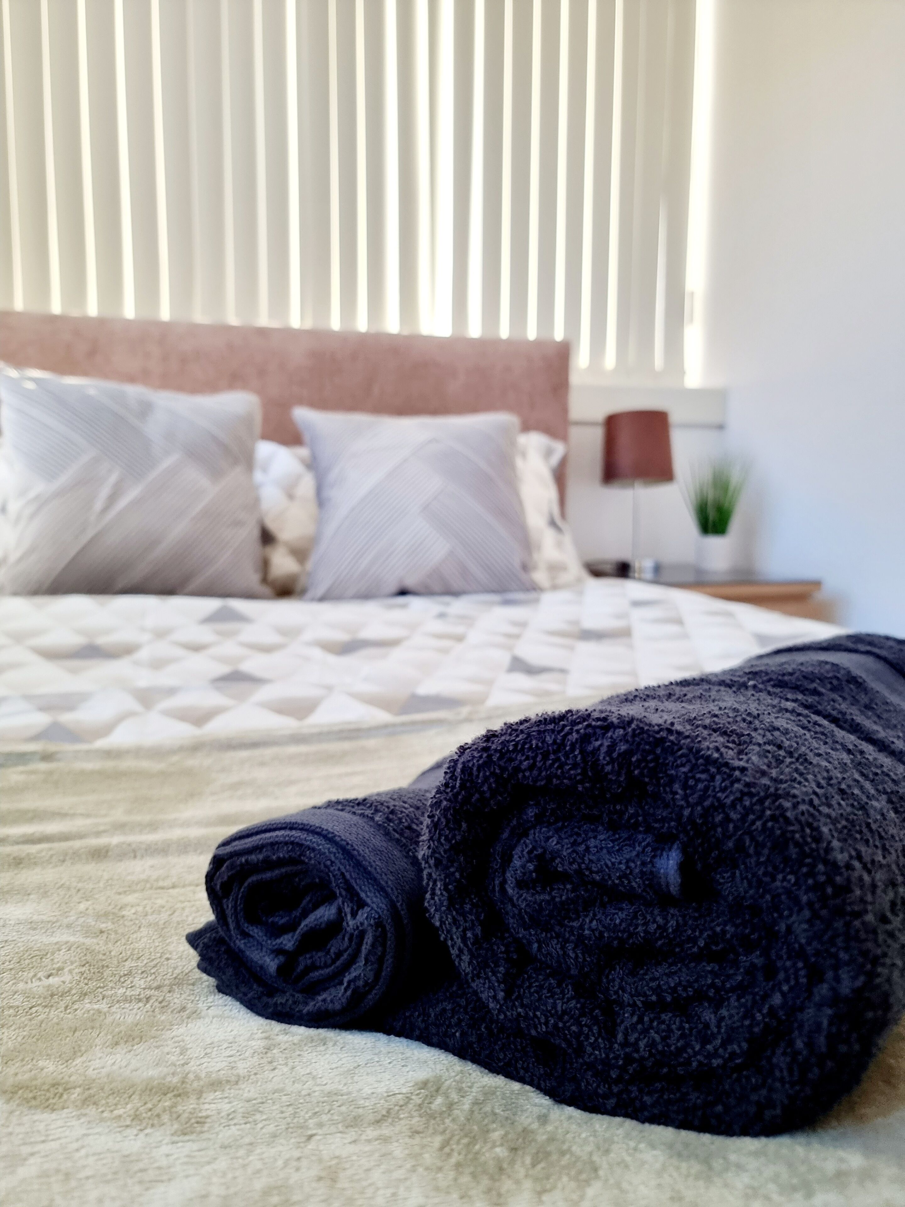 A neatly made bed with a rolled-up dark towel in the foreground, a patterned bedspread, and a headboard with pillows.
