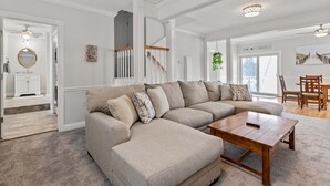 Un salón amplio con un sofá seccional beige grande, una mesa de centro de madera y un comedor con sillas de madera. Una escalera con barandilla de madera conduce a un nivel superior.