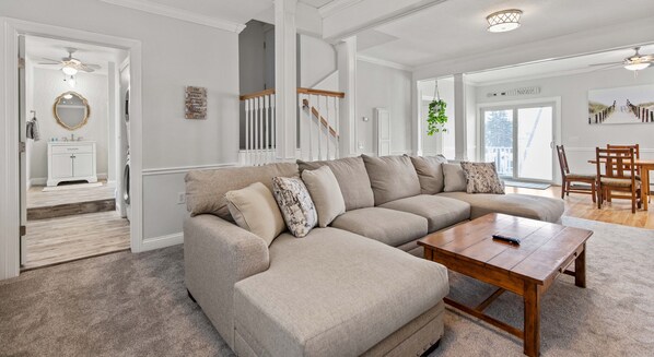 A living room with a beige sectional sofa, a wooden coffee table, and a dining area with wooden chairs.