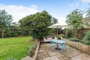 A patio with a table and chairs, a white umbrella, and a garden with a variety of plants and trees. - Barton Cottage Bed and Breakfast (Dorchester)