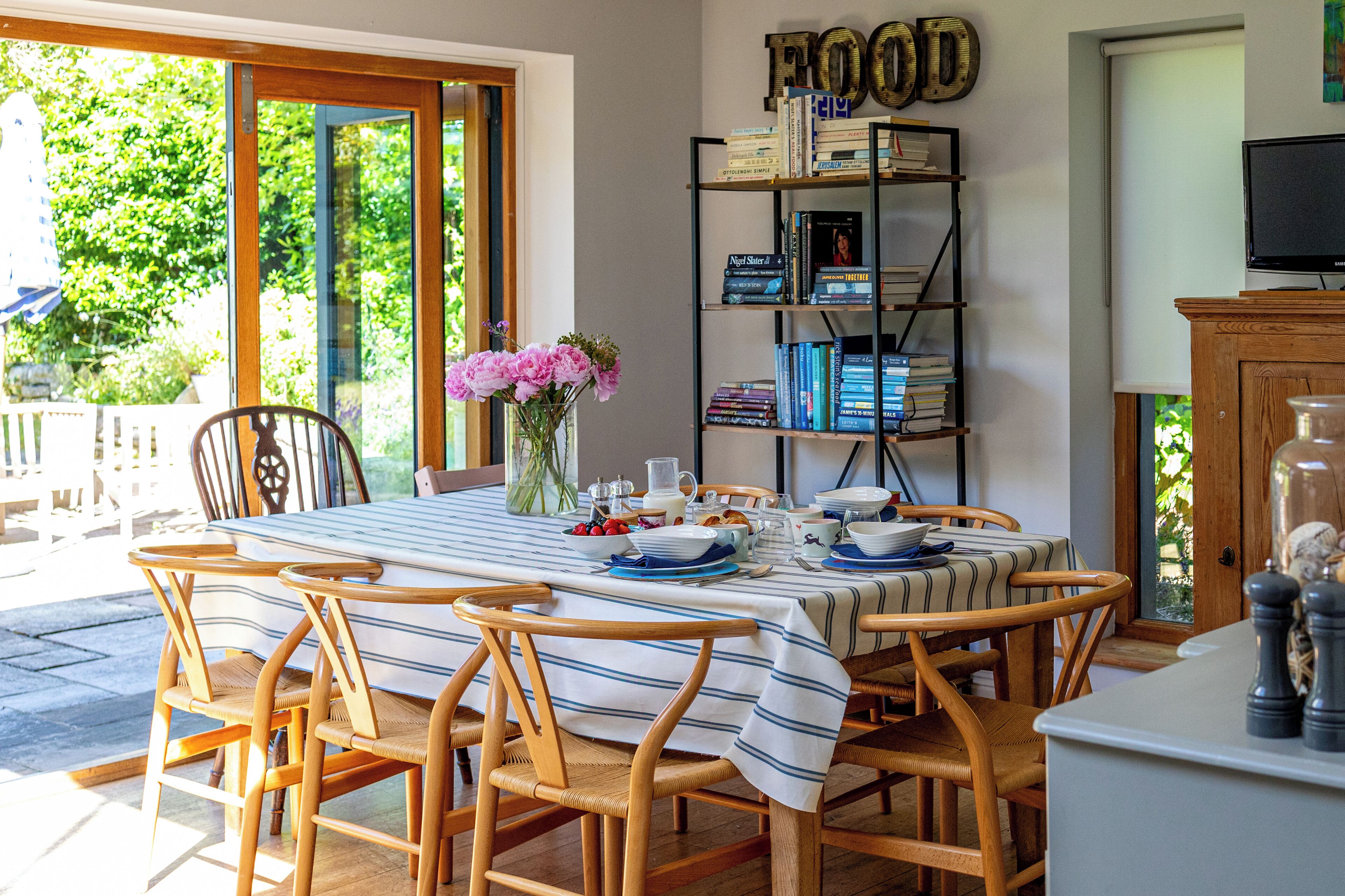 Un coin repas avec une table dressée pour quatre personnes, entourée de chaises en osier, une bibliothèque remplie de livres et un téléviseur posé sur un meuble.