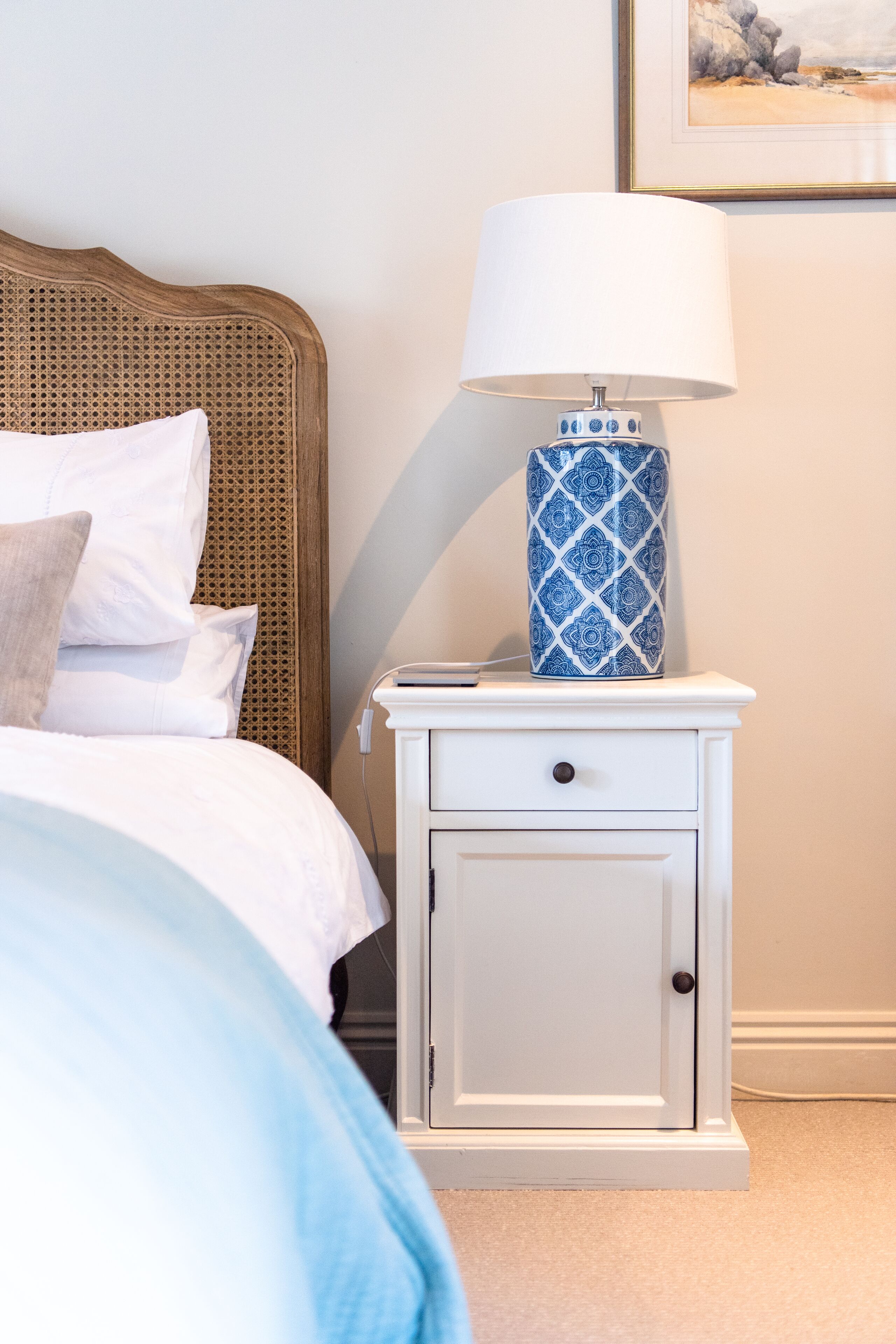 Un lit bien fait, avec du linge de lit blanc et une tête de lit marron, une table de chevet blanche avec une lampe à motifs bleus, et un tableau encadré accroché au mur.