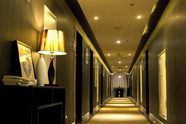 A hotel hallway with a reception desk, a lamp, framed pictures, and doors leading to rooms.