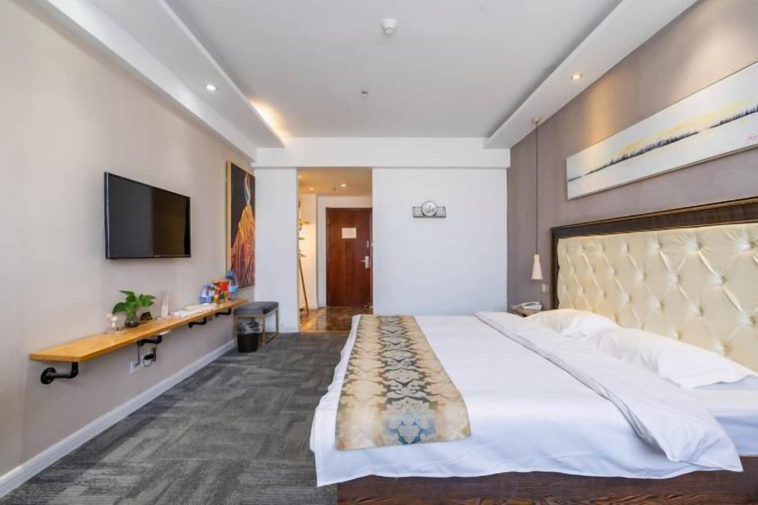 Habitación de hotel con una cama, televisión, estante con objetos, un cuadro y una puerta que da a otra habitación.