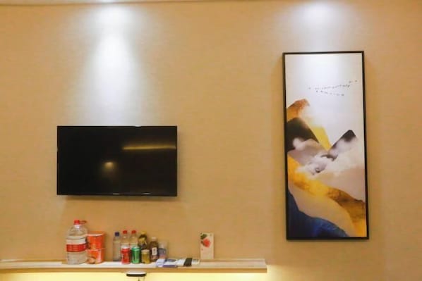 A flat-screen TV mounted on a wall above a shelf with various items including a water bottle, cans, and a small box. - Loft Hotel (Changyi)