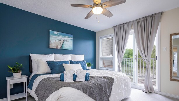 A bedroom with a bed, a nightstand, a ceiling fan, a window with curtains, and a view of greenery outside. - Beach1 Waterfront Stunning Views (Wasaga Beach)