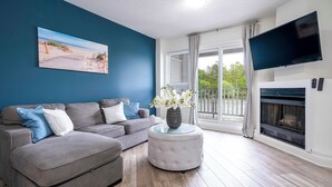 A living room with a grey sofa, a round coffee table with a vase of flowers, a flat-screen TV mounted on the wall, and a fireplace. - Beach1 Waterfront Stunning Views (Wasaga Beach)