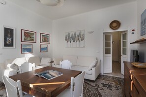 Un salon avec une table en bois, des chaises blanches, un canapé blanc et un tapis à motifs. Des tableaux encadrés ornent le mur, et une porte donne accès à une autre pièce.