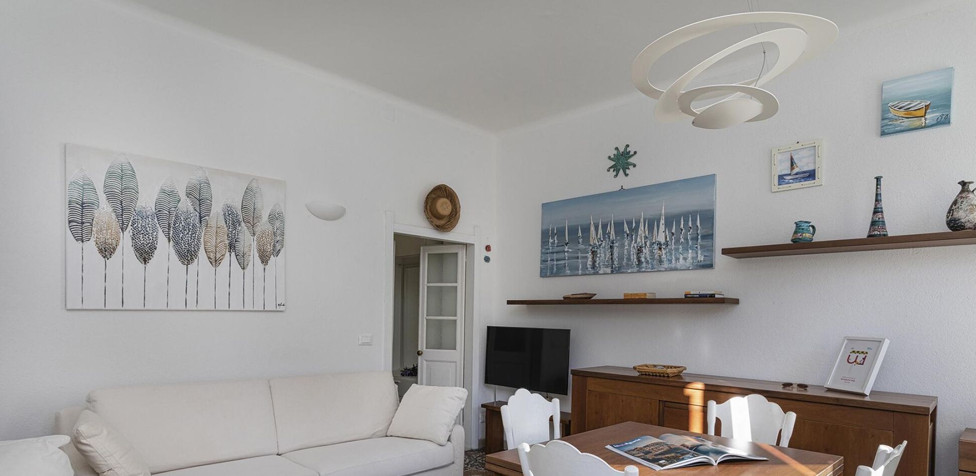 Een woonkamer met een witte bank, een eettafel met stoelen, een televisie en kunstwerken aan de muur.