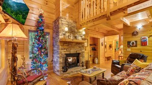 Ein gemütliches Blockhaus-Interieur mit Kamin aus Stein, einem Weihnachtsbaum, Holzmöbeln und einem Schild mit der Aufschrift 'The Mountains Are Calling'.