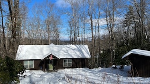 A wooden cabin with a snow-covered roof, surrounded by trees and a clear sky.