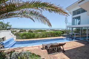 A modern outdoor pool area with a view of the ocean, a patio with seating, and a glass-enclosed structure.