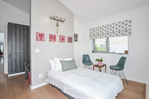 A bedroom with a bed, two chairs, a small table with a plant, a window with heart-patterned curtains, and a wardrobe.