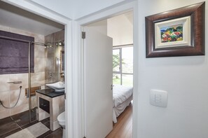 A modern bathroom with a glass shower, a sink, and a toilet. There is a door leading to a bedroom with a bed and a window.