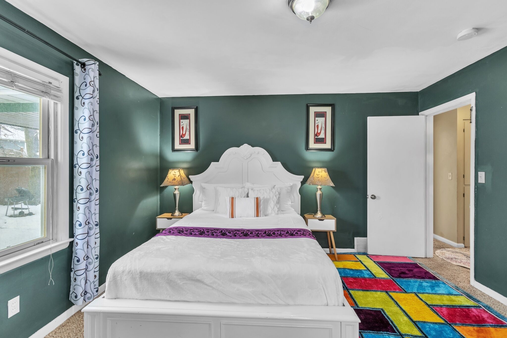 A bedroom with a white bed, two bedside lamps, a colorful rug, and a window with a curtain.