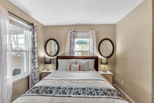 Un dormitorio con cama, dos mesitas de noche, dos espejos en la pared, una ventana con cortinas y vistas al exterior.
