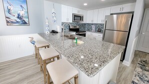 A modern kitchen with white cabinetry, granite countertops, stainless steel appliances, and a central island with bar stools. - 510 South Hampton 2 Bedroom Condo (Myrtle Beach)