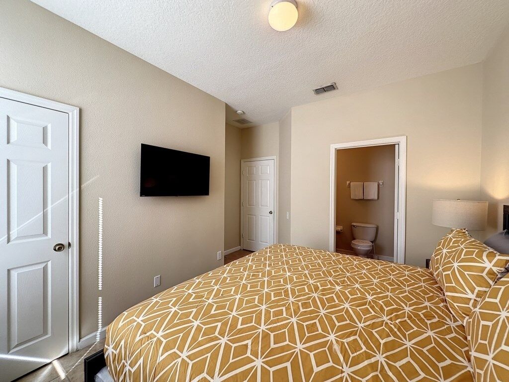 A bedroom with a large bed, a television mounted on the wall, a door, and a bathroom visible through an open door.