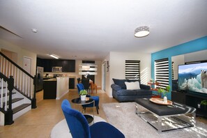 Uma sala de estar moderna com sofá, mesa de centro e TV. É possível ver uma cozinha ao fundo, com frigorífico e micro-ondas.