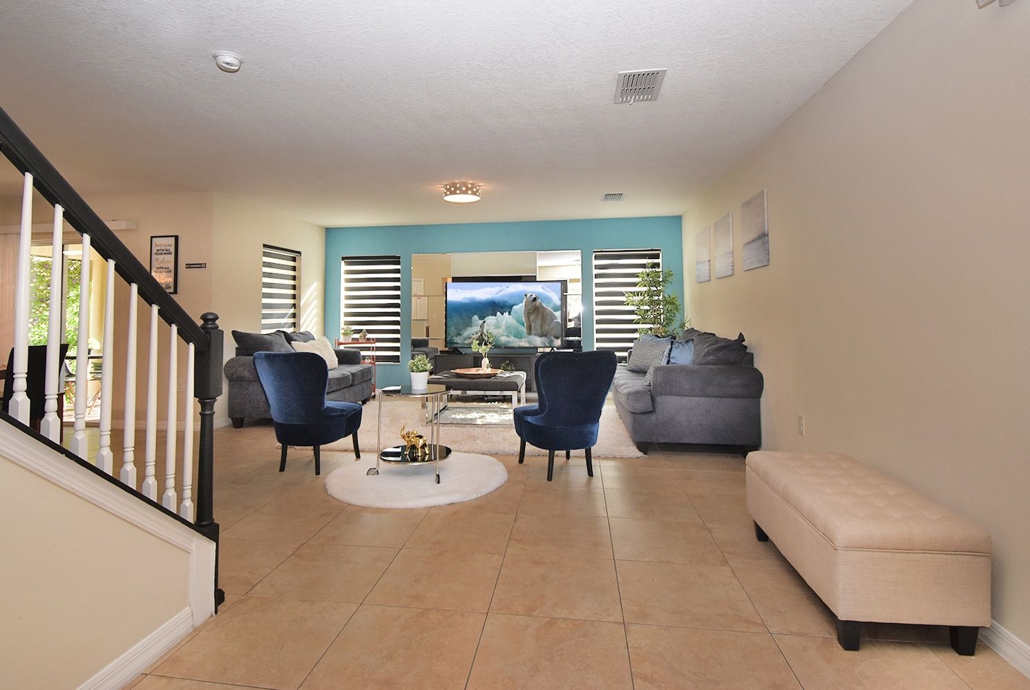 A spacious living room with a staircase, a flat-screen TV, and a large window with blinds.