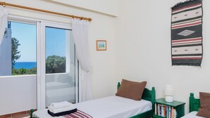 Une chambre avec deux lits, un balcon offrant une vue, une table de chevet verte avec des livres, et un tableau encadré au mur.