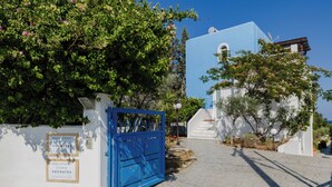 Une porte bleue mène à un bâtiment blanc surmonté d’un toit bleu, le tout étant entouré de verdure et se dressant sous un ciel dégagé.