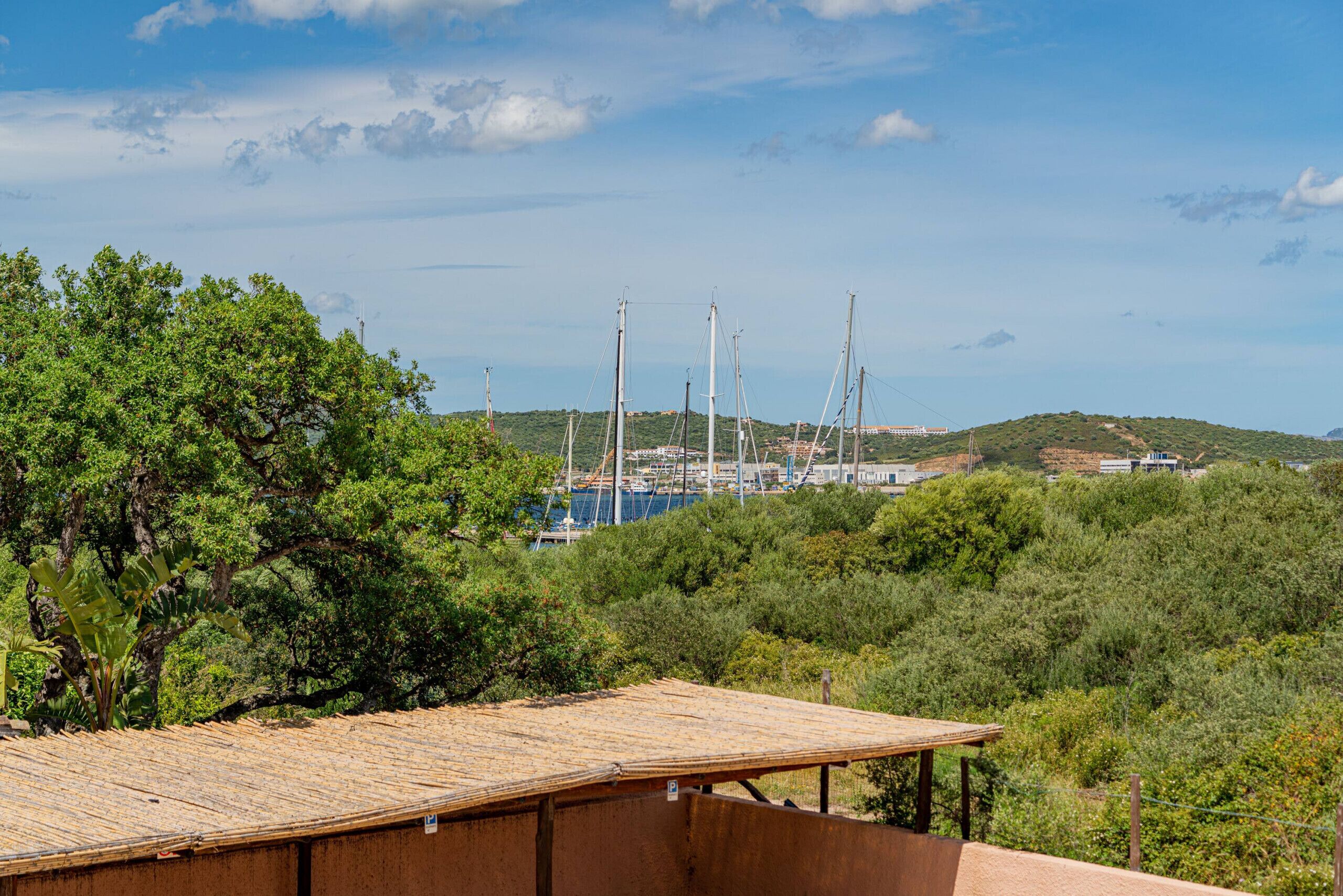 A coastal view with a wooden structure in the foreground, a marina with sailboats in the background, and hills with sparse vegetation.