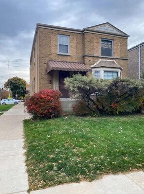 A two-story brick building with a front porch, a small front yard with grass and shrubs, and a visible sidewalk. - Spacious 3BR Home w Parking. Perfect for Families & Longer Stays (Chicago)