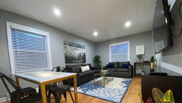 En moderne stue med sofa, sofabord, spisebord med stole og et stort vindue med persienner.