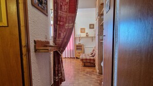 Un couloir étroit au sol carrelé, une porte en bois sur la gauche et, au fond, un lit recouvert d’une couverture à carreaux.