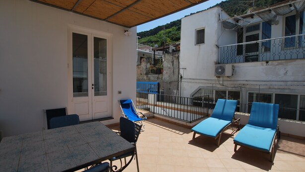 A covered outdoor area with a table and chairs, a balcony with blue lounge chairs, and a white building with a balcony. - Casa Al Mar Canneto Via Mazzini (Lipari)