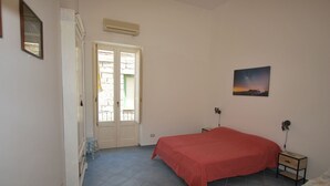 A bedroom with a bed, a nightstand, a lamp, a painting, and a door leading to an outdoor area. - Casa Al Mar Canneto Via Mazzini (Lipari)