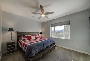 Un dormitorio con cama, dos mesitas de noche, ventilador de techo, ventana con vistas a la montaña y piso alfombrado.
