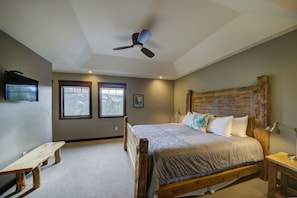 A bedroom with a wooden bed frame, a ceiling fan, two windows, a bench, and a TV mounted on the wall.