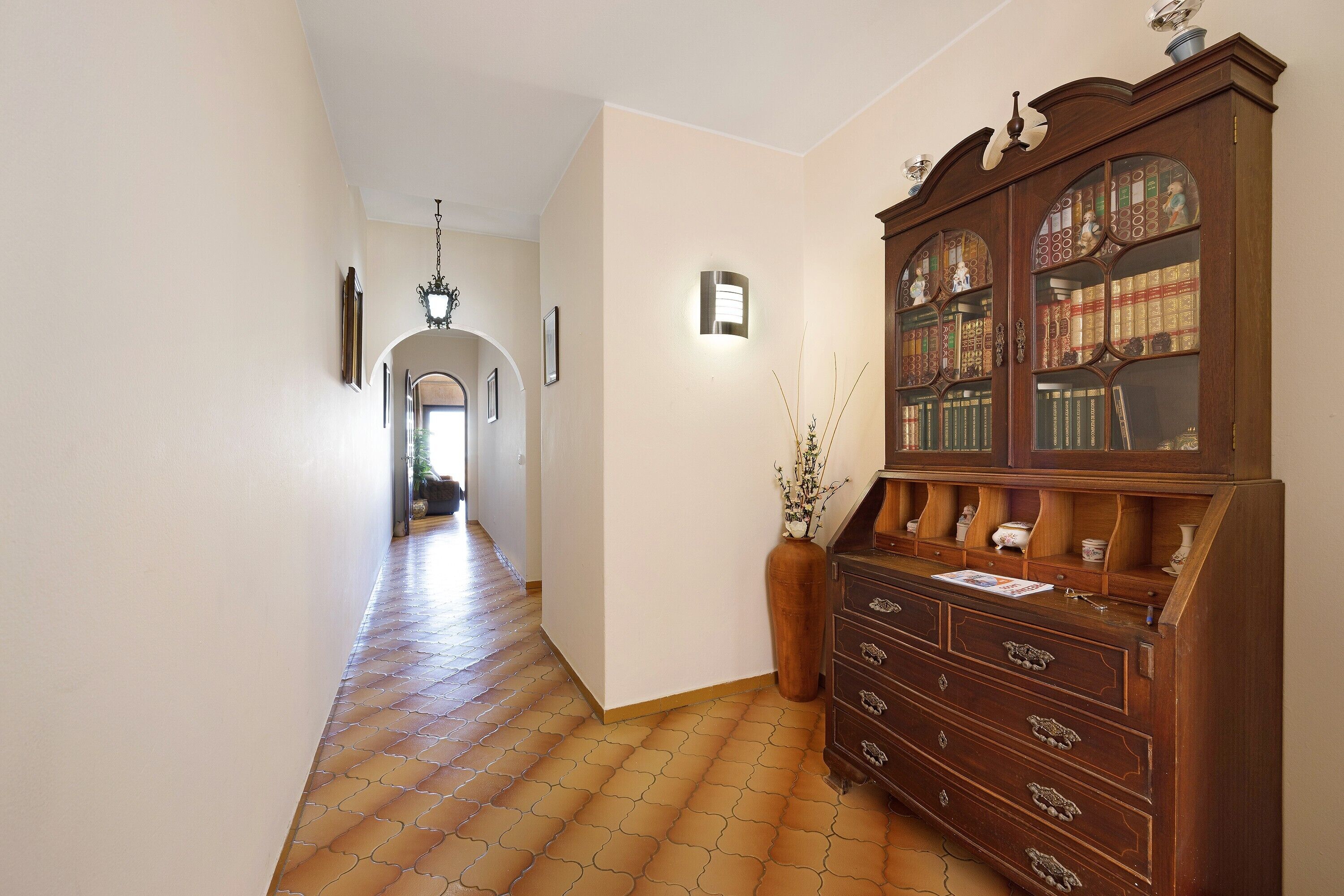 A hallway with a wooden cabinet, a vase with plants, and a tiled floor.